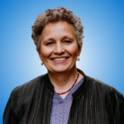 A woman with short curly grey hair, wearing glasses on her head, earrings, a dark striped jacket, and a lavender blouse, smiles in front of a blue gradient background—perfectly representing engaging workshop hosts or panellists.