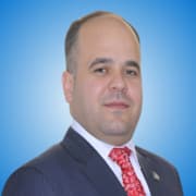A man with short dark hair and a receding hairline, dressed in a dark pinstripe suit, white shirt, and red patterned tie, poses against a blue gradient background—an experienced speaker often seen among distinguished panellists.