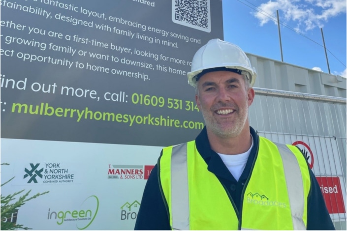A smiling man in a white hard hat and yellow safety vest stands in front of a construction site sign advertising Mulberry Homes Yorkshire, with York & North Yorkshire Combined Authority logos and a metal fence in the background.
