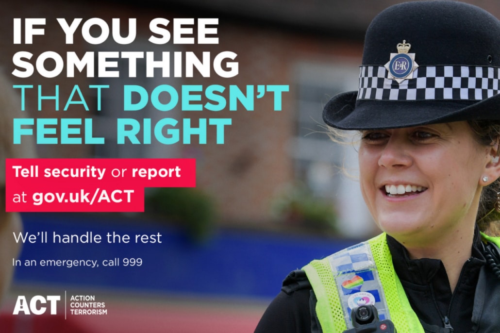 A smiling British police officer in uniform stands outdoors. Text reads: “If you see something that doesn’t feel right, tell security or report at gov.uk/ACT. We’ll handle the rest. In an emergency, call 999.”.