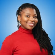 A woman with long, dark, plaited hair wearing a red polo neck smiles against a blue background, embodying the confidence of distinguished speakers.