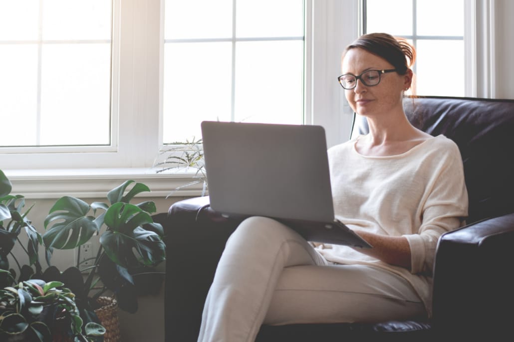 A woman wearing glasses sits on a dark armchair by a window, working on bid writing on her laptop. She is dressed in light-coloured clothes and surrounded by green potted plants. Natural light fills the cosy room.