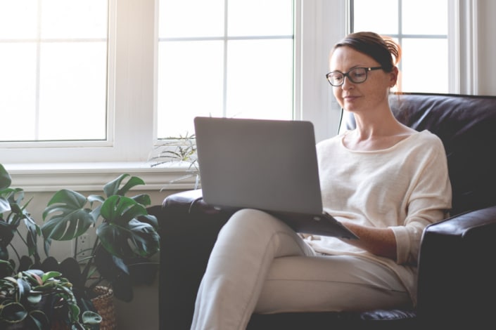 A woman wearing glasses sits on a dark armchair by a window, working on bid writing on her laptop. She is dressed in light-coloured clothes and surrounded by green potted plants. Natural light fills the cosy room.