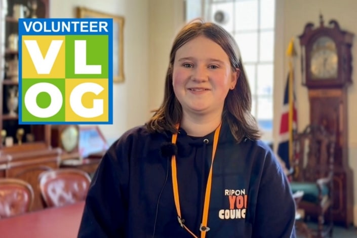 A young person wearing a Ripon Youth Council hoodie smiles at the camera in a formal room, sharing their inspiring journey. A Volunteer VLOG sign with colourful blocks is displayed in the top left corner.
