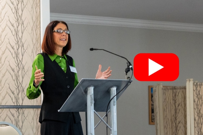 A woman wearing glasses and a green shirt speaks at a lectern with a microphone, delivering an inspiring morning address. A large red YouTube play button icon appears to the right of her, marking the halfway point of the Create 26 event.