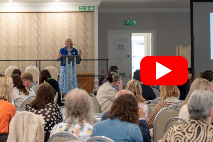 A woman shares her ideas with energy as she speaks at a lectern to a seated audience in a conference room; a large red YouTube play button icon is overlaid on the right side of the image.