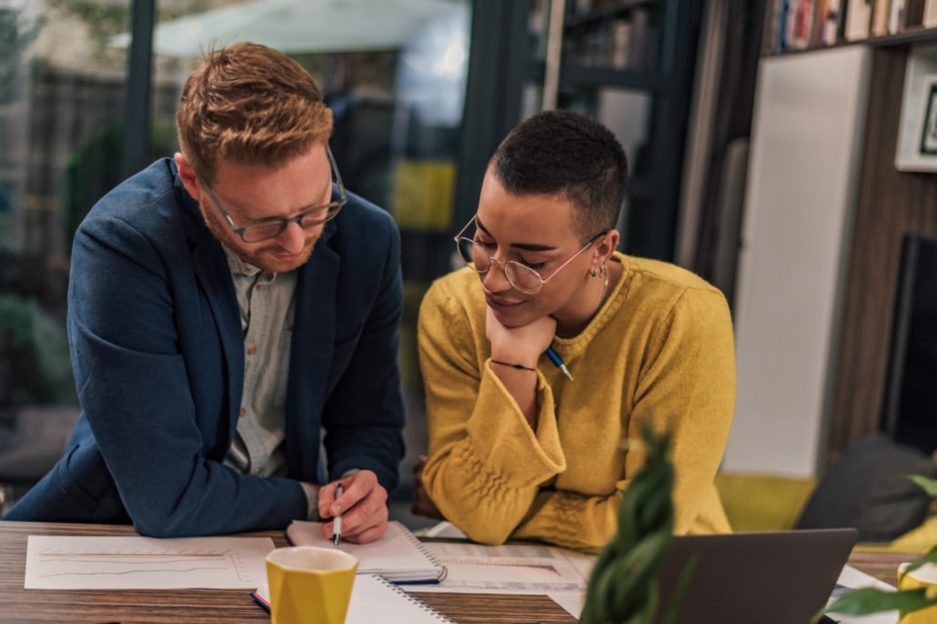 Two people sit at a workplace table, reviewing documents together. One points at a paper with a pen whilst the other looks on thoughtfully. A laptop, notebook, coffee mug, and bookshelves are in the background.
