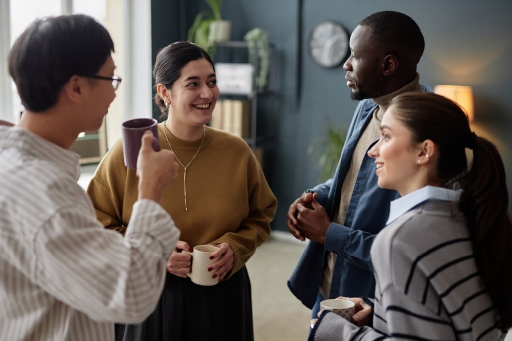 Four people stand in a circle indoors, casually talking and holding mugs. In a friendly office setting with plants and shelves, they discuss barriers to employment whilst appearing engaged and supportive of each other's experiences.