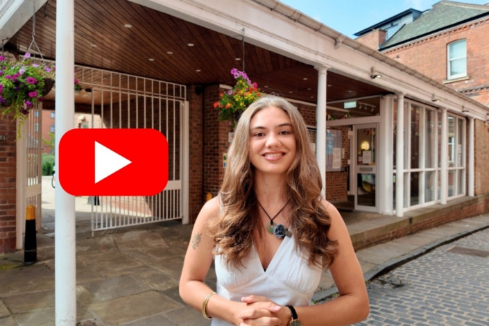 A young woman with long hair in a white dress smiles outside a brick building, where hanging flowers decorate the entrance. A large red YouTube play button logo is on the left side of the image, capturing the lively spirit of a community event such as the Community Climate Conference.