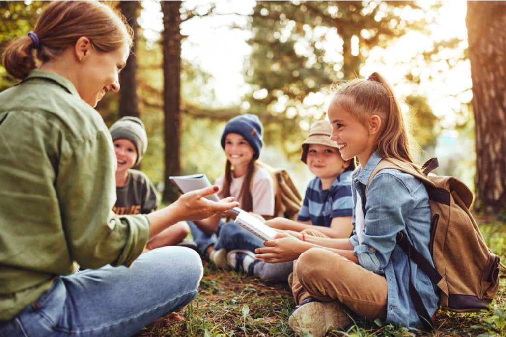 A woman and four children sit in a circle on grass in a North Yorkshire forest, smiling and talking together. The children wear rucksacks, hats, and casual clothes, basking in the warm sunlight filtering through the trees at one of the BSIL Affiliated Sites.