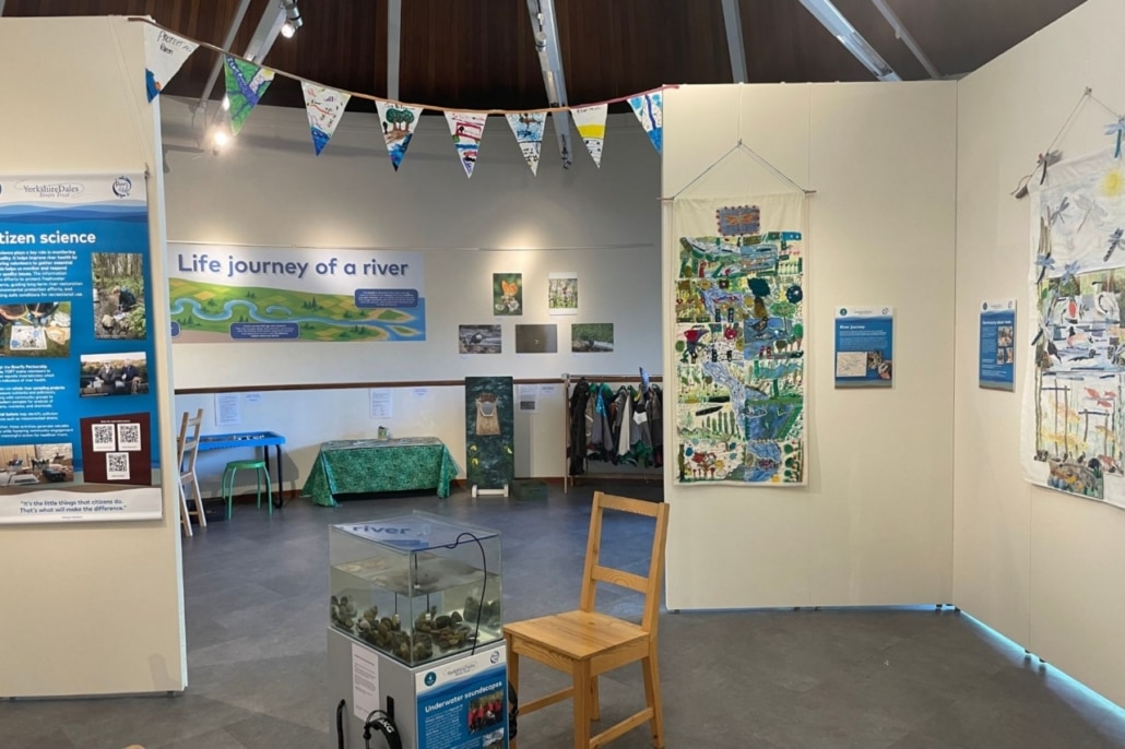 Exhibition room with banners, posters, and quilts about rivers and citizen science. A tank with water and rocks sits by two wooden chairs. Colourful bunting hangs above, and displays cover the Life journey of a river.