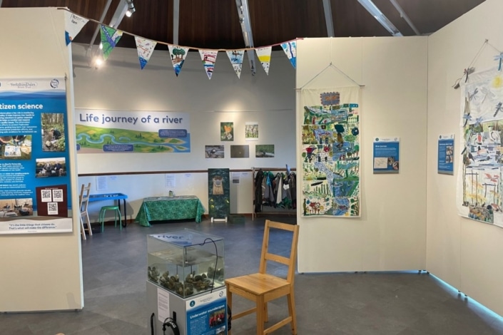 Exhibition room with banners, posters, and quilts about rivers and citizen science. A tank with water and rocks sits by two wooden chairs. Colourful bunting hangs above, and displays cover the Life journey of a river.