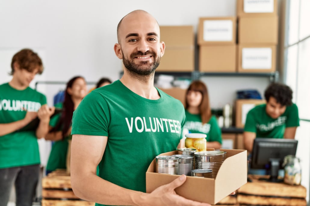 Smiling man in a green Volunteer shirt, recently named Volunteer Leader of the Year, holds a box of tinned food and jars, standing in front of a group of fellow volunteers packing supplies in a donation centre.