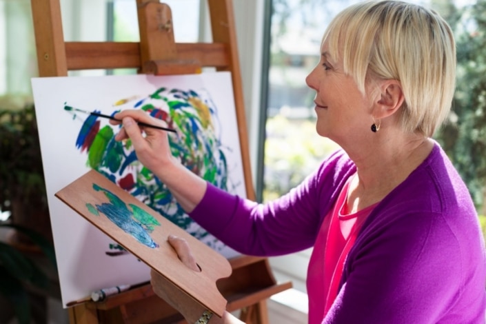 An older woman with short blonde hair and a pink cardigan paints an abstract design at an easel, enjoying social prescribing as she creates art in a bright, sunlit room, holding a palette and brush.