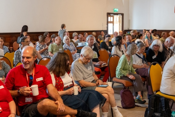 A diverse group of adults sit and chat in a brightly lit conference room with orange chairs and wooden panelling, as they wait for the Community Climate Conference to start. Several people hold mugs or papers, eager to engage in climate action at this inspiring community event.