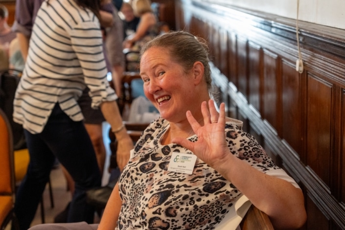A woman with a name badge smiles and waves at the camera while sitting in a room with wooden panelling during the Community Climate Conference. Other people are in the background, creating a warm atmosphere of climate action and community engagement.