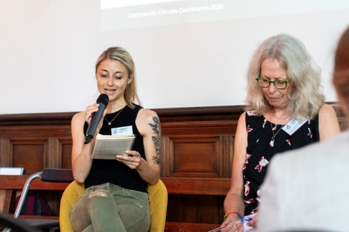 Two women sit side by side in a panel discussion at the Community Climate Conference 2026. One woman speaks into a microphone while reading from a notepad; the other looks down and writes. A screen behind them shows Community Climate Conference 2026, highlighting their commitment to climate action during this important community event.