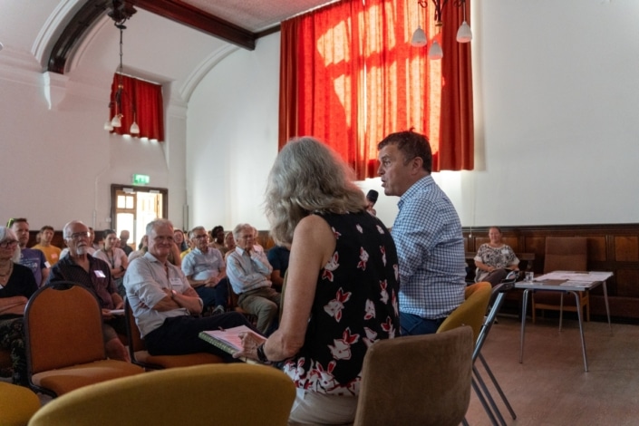 At a Community Climate Conference, a man speaks into a microphone while seated beside a woman in front of an audience in a bright room with red curtains and wooden panelling. People listen attentively, some taking notes, as the discussion centres on community climate action initiatives.