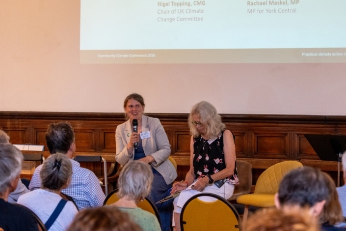 Two women sit at the front of a room speaking to an audience at the Community Climate Conference. One woman holds a microphone whilst the other writes on a notepad, engaging attendees in meaningful discussion about climate action. A projected presentation is displayed on the wall behind them.