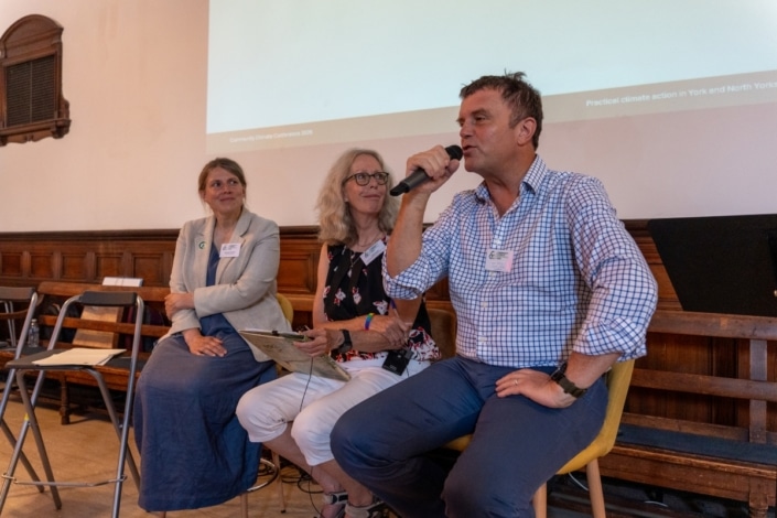 Three people sit on chairs in a panel discussion at the Community Climate Conference. The man on the right holds a microphone and speaks, whilst the two women listen. They all wear name badges and are in a room with wooden panelling and a projection screen, actively engaging in climate action discussions.