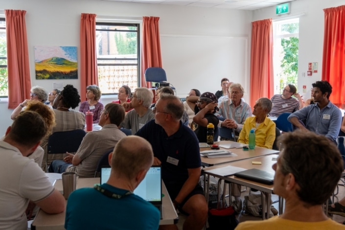 People of various ages sit in a classroom, attentively listening to a speaker out of frame during the Community Climate Conference. Some take notes, and others look towards the front as sunlight streams through windows with orange curtains, creating a vibrant atmosphere for this inspiring community event focused on climate action.