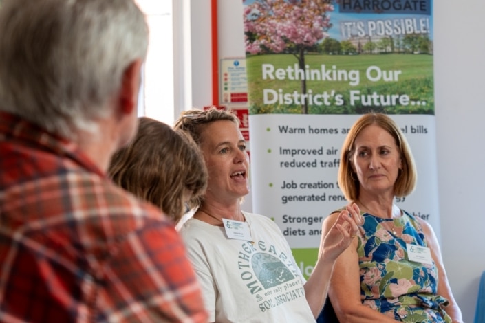 Four adults sit in discussion. One woman speaks, gesturing with her hand. Another woman listens attentively. A poster behind them mentions rethinking the district’s future, with topics like climate and local jobs—highlighting this as a community event focused on climate action and collaboration.