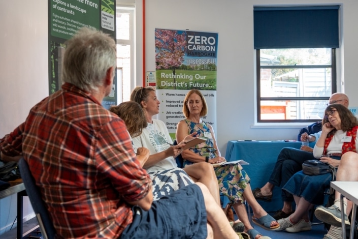 A group of people sit in a room having a discussion, with posters about zero carbon and sustainability on the wall behind them. Some people hold papers and listen attentively, taking part in a Community Climate Conference. Sunlight comes in through a window, highlighting the importance of climate action at this community event.
