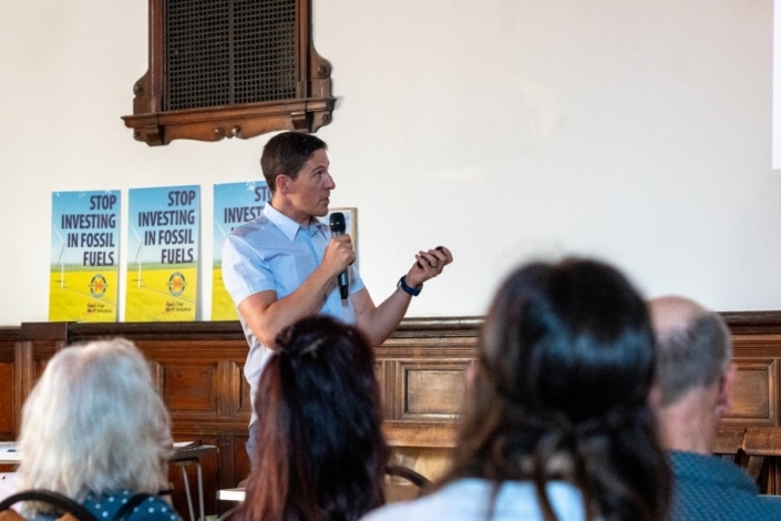 A person speaks into a microphone whilst pointing at a screen, with “Stop Investing in Fossil Fuels” posters visible behind them during the Community Climate Conference. Several people in the audience watch attentively, engaging in this impactful community event dedicated to climate action.