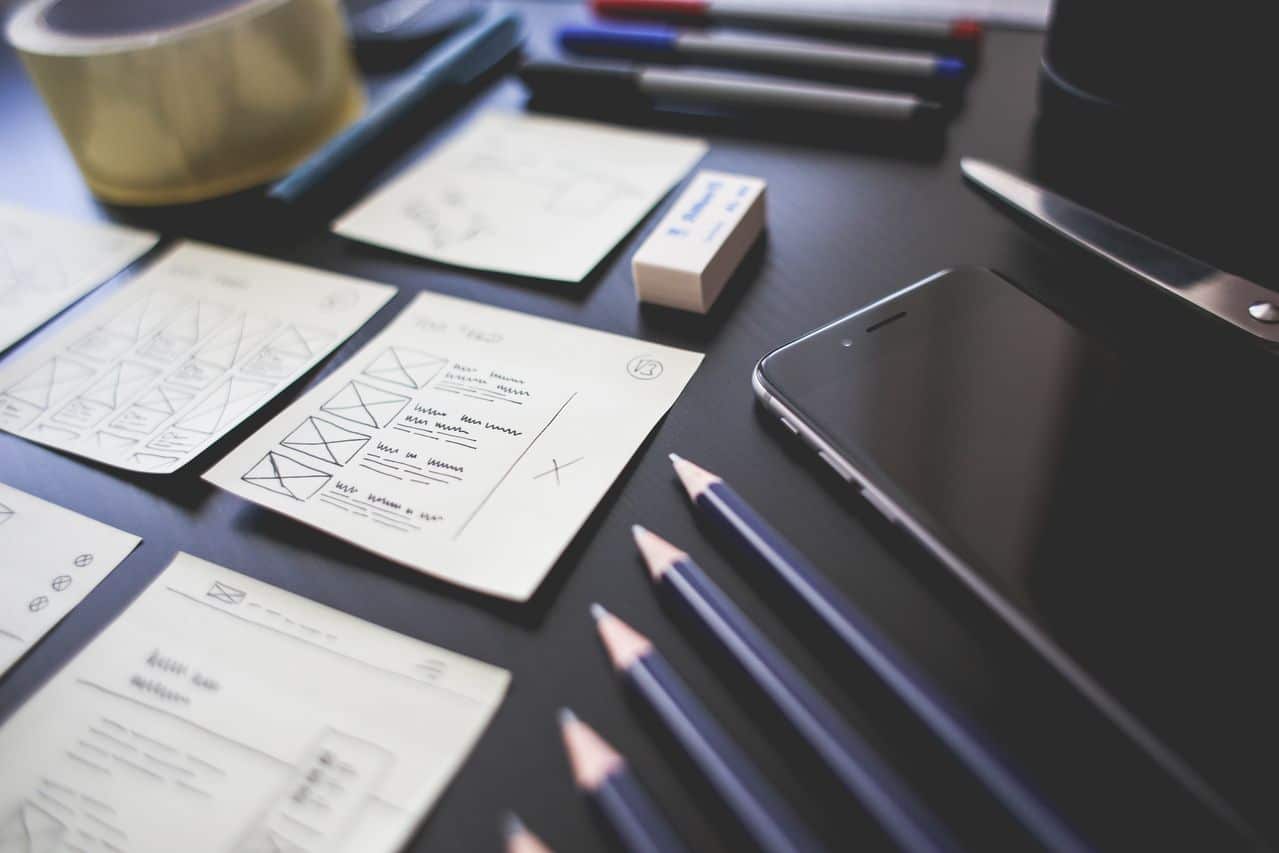 A workspace with pencils, pens, sticky notes with sketches, a rubber, a mobile phone, tape, and scissors arranged neatly on a dark table.