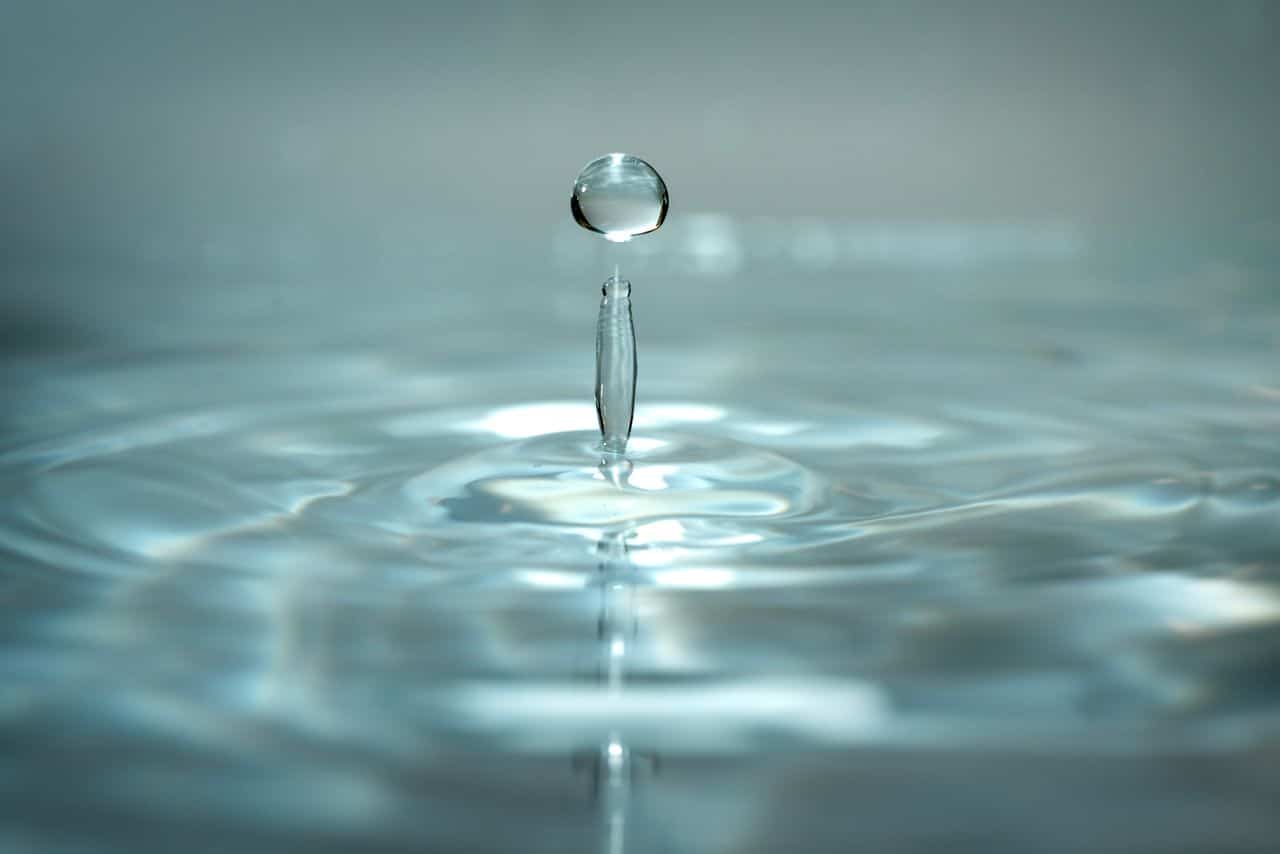 A close-up photo of a water droplet suspended above the surface, causing ripples in the clear water below. The background is softly blurred.