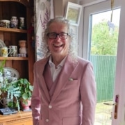 A smiling man with grey curly hair and glasses wears a light pink suit and stands indoors near a window, surrounded by plants and decorative items. His confident presence reflects his experience in leadership and mentoring programmes.