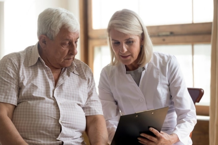 A doctor in a white coat shows a clipboard to an older man as they sit together indoors, discussing medical information and the future of adult social care near a window.
