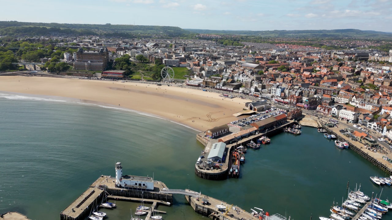Aerial view of a coastal town with a sandy beach, a pier with boats, a ferris wheel near the shore, and a mix of buildings and green hills in the background under a partly cloudy sky.