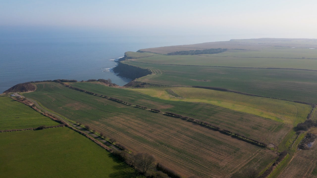 Aerial view of green coastal farmland with hedgerows, fields, and a rocky shoreline meeting the calm sea under a hazy sky.