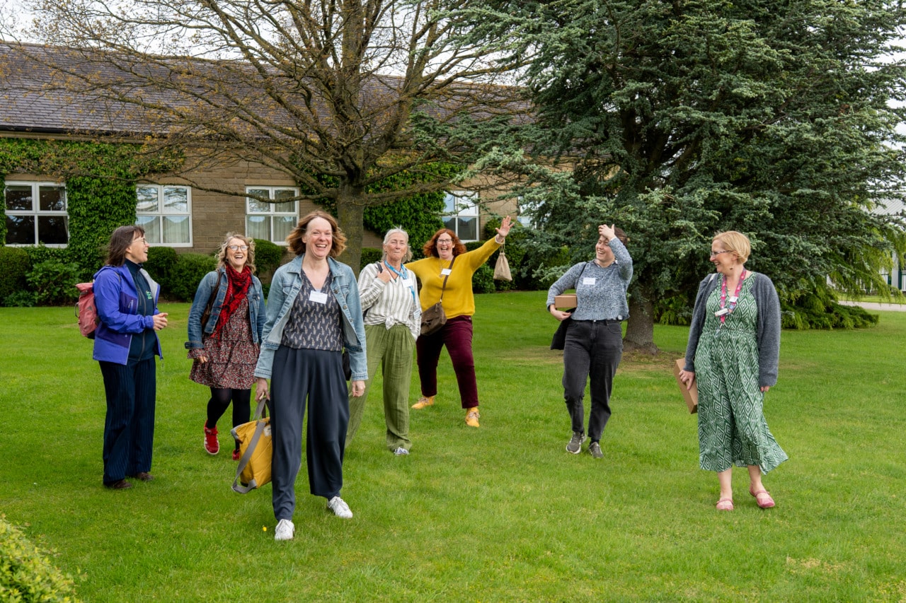 Seven adults stand and walk on green grass, smiling and laughing together. A stone building with ivy and large trees are in the background, suggesting a relaxed, outdoor gathering.