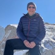 A person wearing sunglasses, a blue winter jacket, and a beanie is smiling whilst sitting on a snowy rock with mountains and a clear blue sky in the background, embodying the spirit of leadership and adventure found in many successful leadership programmes.
