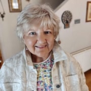 An older woman with short, light hair smiles indoors. She wears a patterned shirt and light jacket, embodying a sense of leadership and warmth. The background shows home decor, a radiator, and framed pictures on the wall, suggesting an inviting space—perfect for meaningful mentoring conversations.