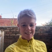 Person with short fair hair wearing a bright yellow jacket and wireless earphones, smiling at the camera outdoors. There is a brick wall and blue sky in the background, capturing a confident moment during their participation in a Leadership Mentoring Programme.