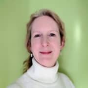 A person with light skin and light brown hair, wearing a white polo-neck jumper and small earrings, smiles softly in front of a plain light green background, embodying the approachable confidence often seen in leadership and mentoring roles.
