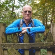 A man wearing sunglasses and a blue jacket leans on a wooden fence in a forested area, with trees and foliage in the background, embodying the reflective spirit often found in those participating in a Leadership Mentoring programme.
