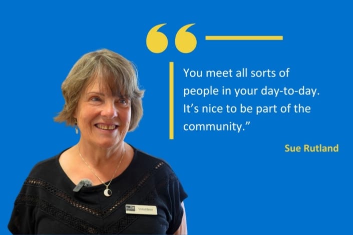 Woman with short hair and a name badge smiles in front of a blue background with a quotation about community involvement, attributed to Sue Rutland, in white and yellow text—highlighting Sue’s story of volunteering at The Forum.