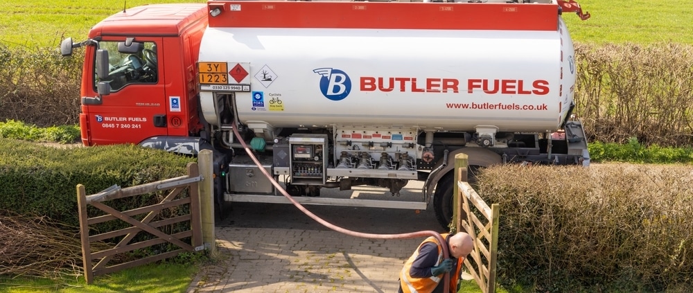 A Butler Fuels tanker lorry is parked on a drive, delivering fuel to rural families. A worker in an orange safety vest walks on a stone path, holding a hose connected to the lorry. The background shows green fields and hedges.