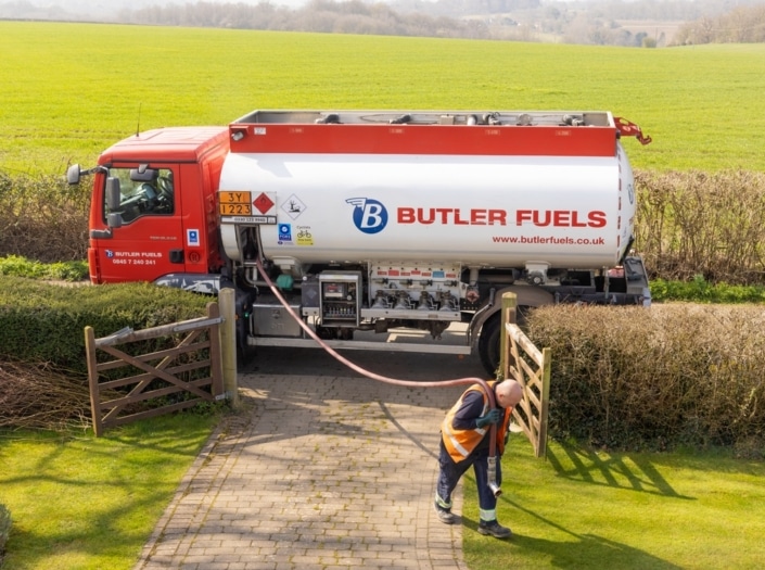 A Butler Fuels tanker lorry is parked on a drive, delivering fuel to rural families. A worker in an orange safety vest walks on a stone path, holding a hose connected to the lorry. The background shows green fields and hedges.