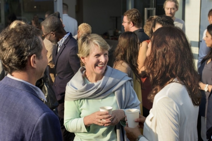 A woman smiling and holding a paper cup whilst chatting with others at an outdoor gathering.