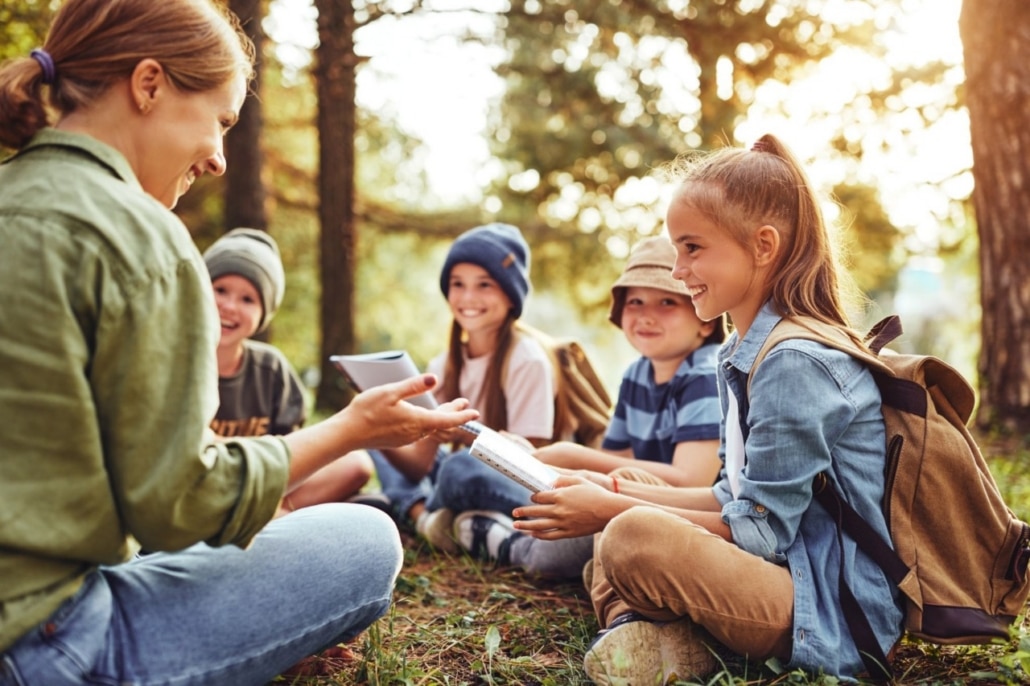 A teacher sits in a circle outdoors, talking to four pupils who are holding exercise books.