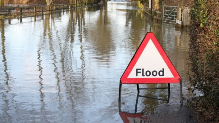 A triangular red and white Flood road sign stands in a flooded road, serving as a reminder to be prepared.
