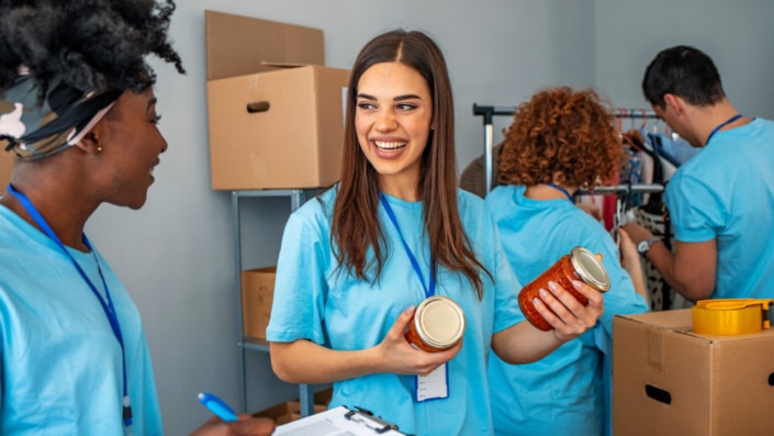 Four youth volunteers in light blue shirts work in a donation centre, with one woman in the foreground smiling while holding a jar of food.