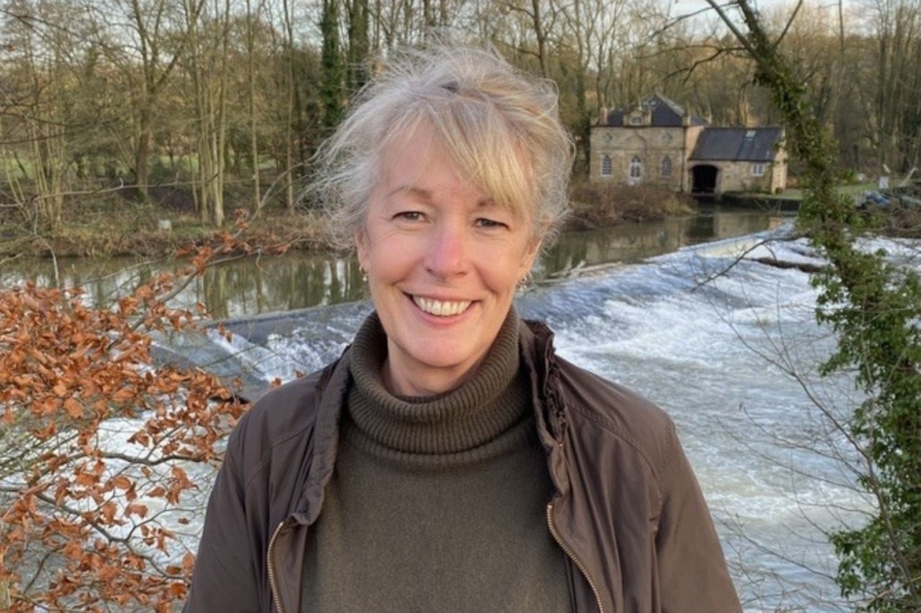 A smiling woman stands in front of a river weir and a stone building, representing local community engagement.