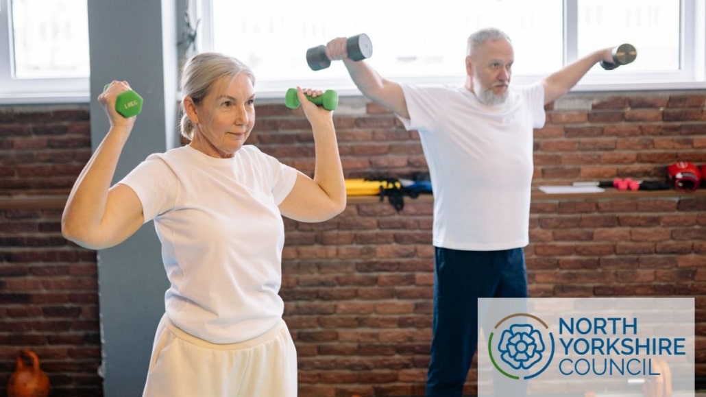 An older man and woman performing overhead dumbbell presses in a gym as part of a programme supporting healthy ageing, with the North Yorkshire Council logo in the corner.
