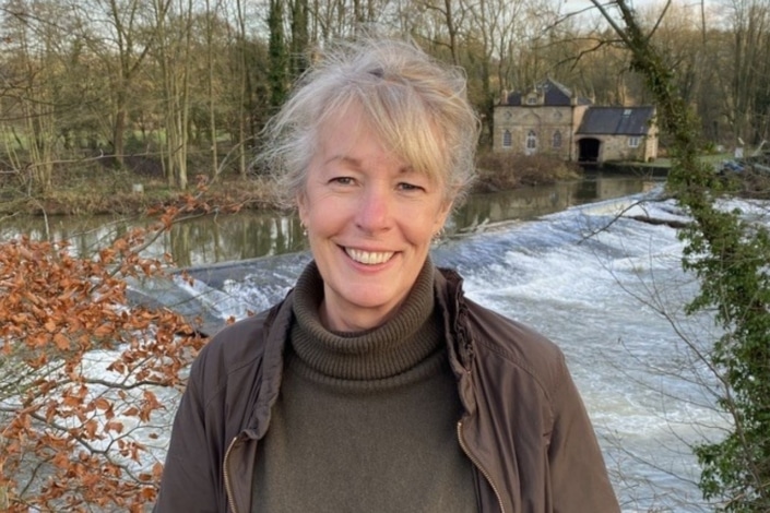 A smiling woman stands in front of a river weir and a stone building, representing local community engagement.
